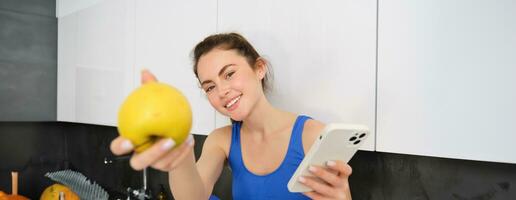 Portrait of beautiful, smiling young fitness woman, offering you an apple, eating healthy snack, holding a fruit, sitting in kitchen photo