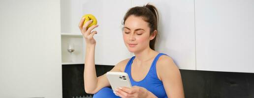 retrato de morena aptitud mujer, comiendo un manzana, participación teléfono inteligente, utilizando móvil teléfono aplicación mientras teniendo sano Fruta bocadillo en cocina foto