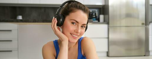 Close up portrait of beautiful brunette girl in sportswear, sits in wireless headphones and listens music, while doing workout training at home photo