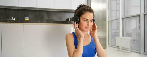 Image of fitness girl doing sports at home, puts on wireless headphones for music during workout training session, wearing blue sportsbra photo