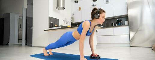 retrato de mujer en inalámbrico auriculares, haciendo rutina de ejercicio desde hogar, en pie en tablón con Deportes equipo, escuchando música, mirando confidente foto