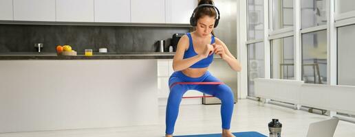 retrato de activo y sano joven mujer, haciendo sentadillas con resistencia banda, rutina de ejercicio a hogar, escuchando música en inalámbrico auriculares foto