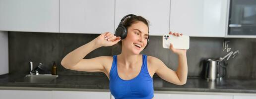 retrato de hermosa aptitud bloguero, mujer en auriculares, escuchando música y bailando en cocina, vistiendo azul polainas y sujetador deportivo foto