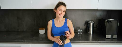 retrato de ajuste y sano sonriente mujer, vistiendo aptitud ropa de deporte, en pie en cocina con agua botella y mirando contento foto