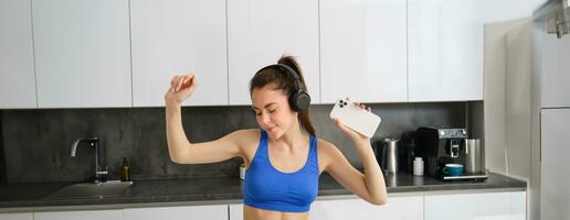 retrato de hermosa aptitud bloguero, mujer en auriculares, escuchando música y bailando en cocina, vistiendo azul polainas y sujetador deportivo foto