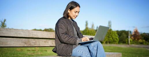 Digital nomad. Portrait of young woman using laptop in park, sitting on bench and working, studying online photo