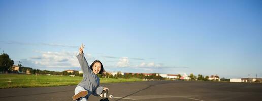 Vertical shot of happy asian skater girl, jumping, standing with skateboard and smiling. Woman skating on longboard and having fun photo