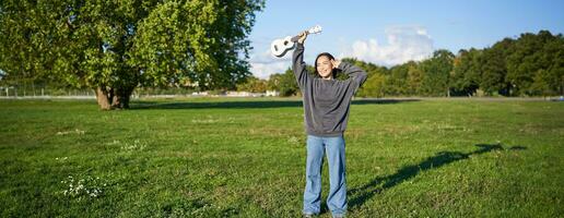 Happy asian girl, musician with ukulele, feeling carefree, enjoying freedom and fresh air outdoors, playing musical instrument photo