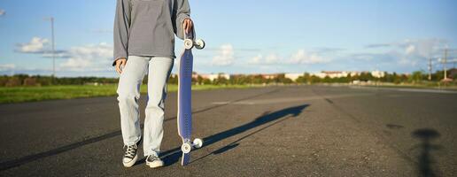 Cropped shot of female body, holding skateboard in hand, walking on road. Young woman carry her longboard, cruiser, skating on the road photo