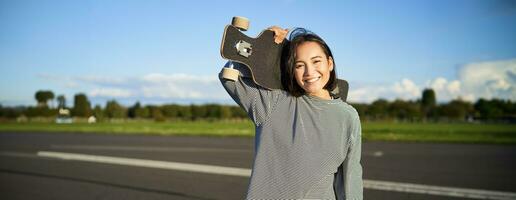 retrato de hermosa joven patinador chica, en pie con longboard y sonriente a cámara. asiático mujer con patineta en pie en la carretera foto