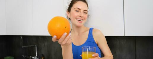 cerca arriba retrato de deportista, aptitud niña participación vaso de Fresco jugo y un naranja en manos, sonriente a cámara, en pie en cocina foto