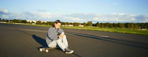 retrato de joven coreano niña sentado en su patineta en camino, mirando a teléfono inteligente, chateando en móvil aplicación foto