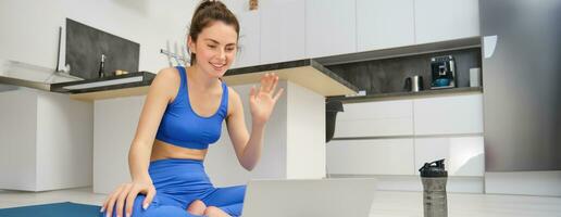 retrato de aptitud instructor, mujer conecta a en línea formación sesión, olas mano a computadora portátil, enseñando yoga rutina de ejercicio desde hogar, sentado en caucho estera foto
