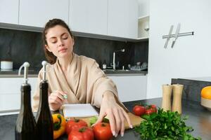 Portrait of woman sitting with groceries, making list for shopping, posing near vegetables, preparing food, cooking in the kitchen photo