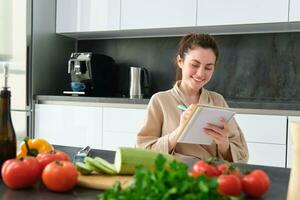 Portrait of happy young woman writes down menu for dinner, sits in the kitchen near vegetables, makes grocery list for shopping, poses in the bathrobe photo