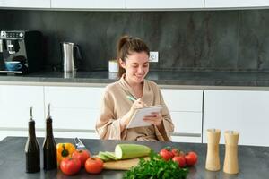 Portrait of beautiful, smiling young woman making list of meals, writing down recipe, sitting in the kitchen with vegetables, doing house errands photo