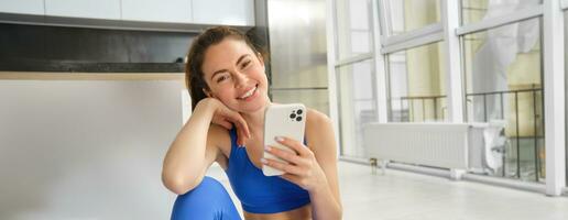 joven mujer con teléfono inteligente lo hace rutina de ejercicio a hogar, utilizando móvil teléfono aplicación para Deportes formación adentro, usa azul sujetador deportivo y polainas foto