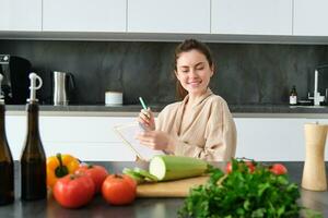 Portrait of woman sitting with groceries, making list for shopping, posing near vegetables, preparing food, cooking in the kitchen photo