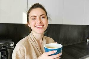 Lifestyle concept. Portrait of happy brunette woman in bathrobe, drinking coffee in the kitchen, having morning cuppa and smiling photo