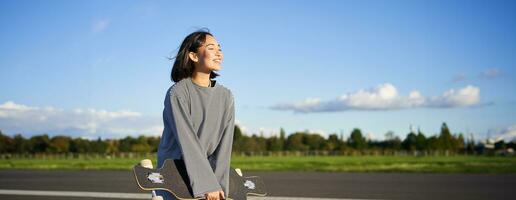 Portrait of asian woman with longboard. Korean girl skating, holding skateboard in hands, posing on road, smiling and looking aside photo