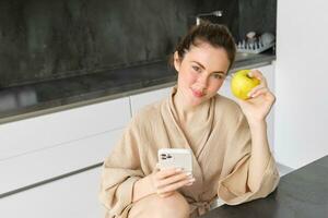 imagen de atractivo joven mujer en bata de baño, se sienta en cocina, come verde manzana y mira a móvil teléfono, usos teléfono inteligente aplicación, pedidos comida en solicitud foto