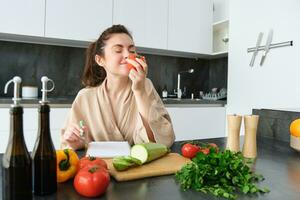 retrato de hermosa joven mujer en el cocina, escritura abajo Cocinando receta, sentado cerca el cortar tablero con vegetales y haciendo tienda de comestibles lista, creando sano vegetariano menú para su familia foto