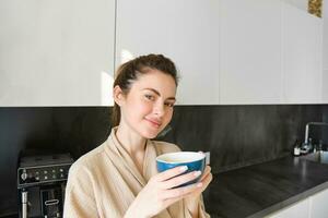 retrato de guapo joven mujer, Bebiendo café en el cocina, disfrutando su Mañana rutina y sonriente a el cámara foto