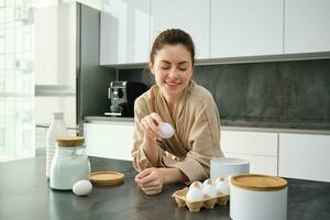 Attractive young cheerful girl baking at the kitchen, making dough, holding recipe book, having ideas photo