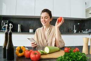 imagen de joven hermosa mujer, participación tomate, sentado en cocina con teléfono inteligente, el cortar tablero y vegetales en encimera, Cocinando alimento, orden comestibles para su receta, utilizando móvil teléfono aplicación foto