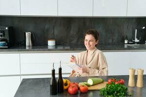 retrato de mujer escritura abajo lista de comestibles, haciendo notas en receta, sentado en cocina cerca verduras, preparando cena menú foto