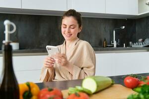 mujer acecho receta en teléfono inteligente, sentado en el cocina con vegetales y el cortar junta, preparando cena, sano ensalada, Cocinando a hogar, vistiendo bata de baño foto