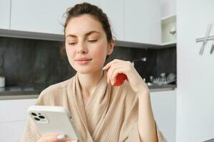 Close up portrait of beautiful smiling woman, holding fresh tomato, sitting in kitchen with smartphone, orders vegetables online, using application to buy groceries, using mobile phone photo