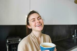 Lifestyle concept. Portrait of happy brunette woman in bathrobe, drinking coffee in the kitchen, having morning cuppa and smiling photo