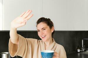 Close up portrait of carefree woman, covers her eyes from sunlight, looking out of window in the kitchen, drinking morning coffee and smiling photo