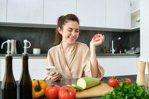 Portrait of young woman ordering groceries on smartphone app, holding tomato, sitting near chopping board with vegetables. Girl looking for recipe online, using mobile phone photo