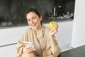 retrato de feliz, hermosa mujer sonriente, comiendo un manzana en el cocina, sentado a hogar en bata de baño, participación teléfono inteligente, utilizando móvil teléfono foto