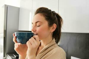 diario rutina y estilo de vida. joven hermosa mujer en bata de baño, en pie en cocina con taza de café, Bebiendo té, sonriente y mirando contento foto