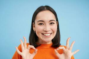 cerca arriba retrato de sonriente hermosa asiático mujer, muestra bueno, Okay bien firmar, aprueba algo, aprueba, da positivo comentario, azul antecedentes foto