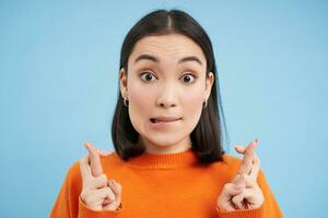 Close up of hopeful asian girl, makes wish, cross fingers, stands over blue background photo