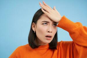 Close up portrait of annoyed korean girl, slaps her forehead, facepalms, rolls eyes irritated, bothered by smth, stands over blue background photo