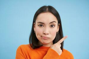 Close up portrait of beautiful modern girl, smirks skeptical, points right, shows banner, stands over blue background photo