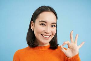 cerca arriba retrato de sonriente hermosa asiático mujer, muestra bueno, Okay bien firmar, aprueba algo, aprueba, da positivo comentario, azul antecedentes foto