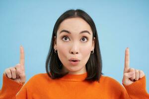Close up of korean woman points up, looks on top with surprised face, amazed by advertisement, stands over blue background photo