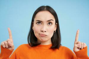 Close up of complicated, puzzled asian girl points up, looks suspicious on top, stands over blue background photo