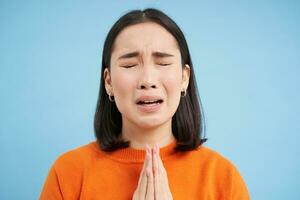 Close up of sad young asian woman, holds hands in pray, begging, pleading for help, stands over blue background photo