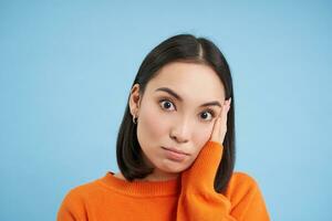 Close up of asian girl looks with shocked face at camera, listens to conversation, stands over blue background photo