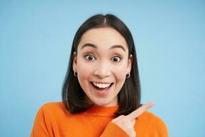 Close up portrait of smiling asian woman, pointing finger right, showing advertisement, copy space banner, standing over blue background photo