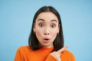 Close up of surprised asian girl, pointing finger right, showing advertisement with curious, intrigued face, standing over blue background photo