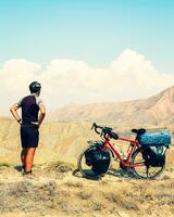 Static back view caucasian male cyclist standing by red touring bicycle looking to scenic mountains background. Active inspirational lifestyle concept photo