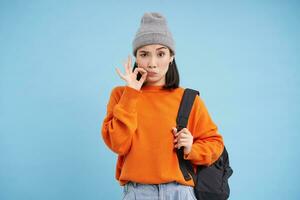 Young asian woman, girl zips her mouth, tells secret, stands over blue studio background photo